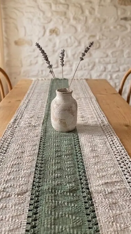 Camino de mesa de macramé verde salvia y crudo sobre una mesa de madera, con un jarrón de cerámica y lavanda