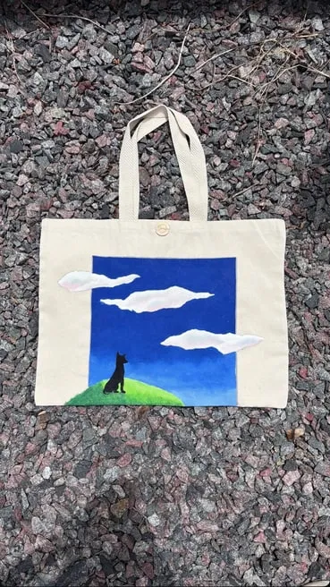 Tote bag de gabardina cruda con un cielo azul, nubes y una figura sobre una colina verde