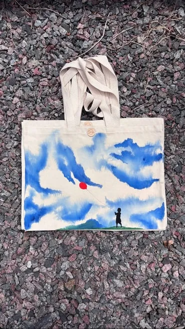 Tote bag de gabardina cruda con nubes azules, un globo rojo y una figura sobre una colina